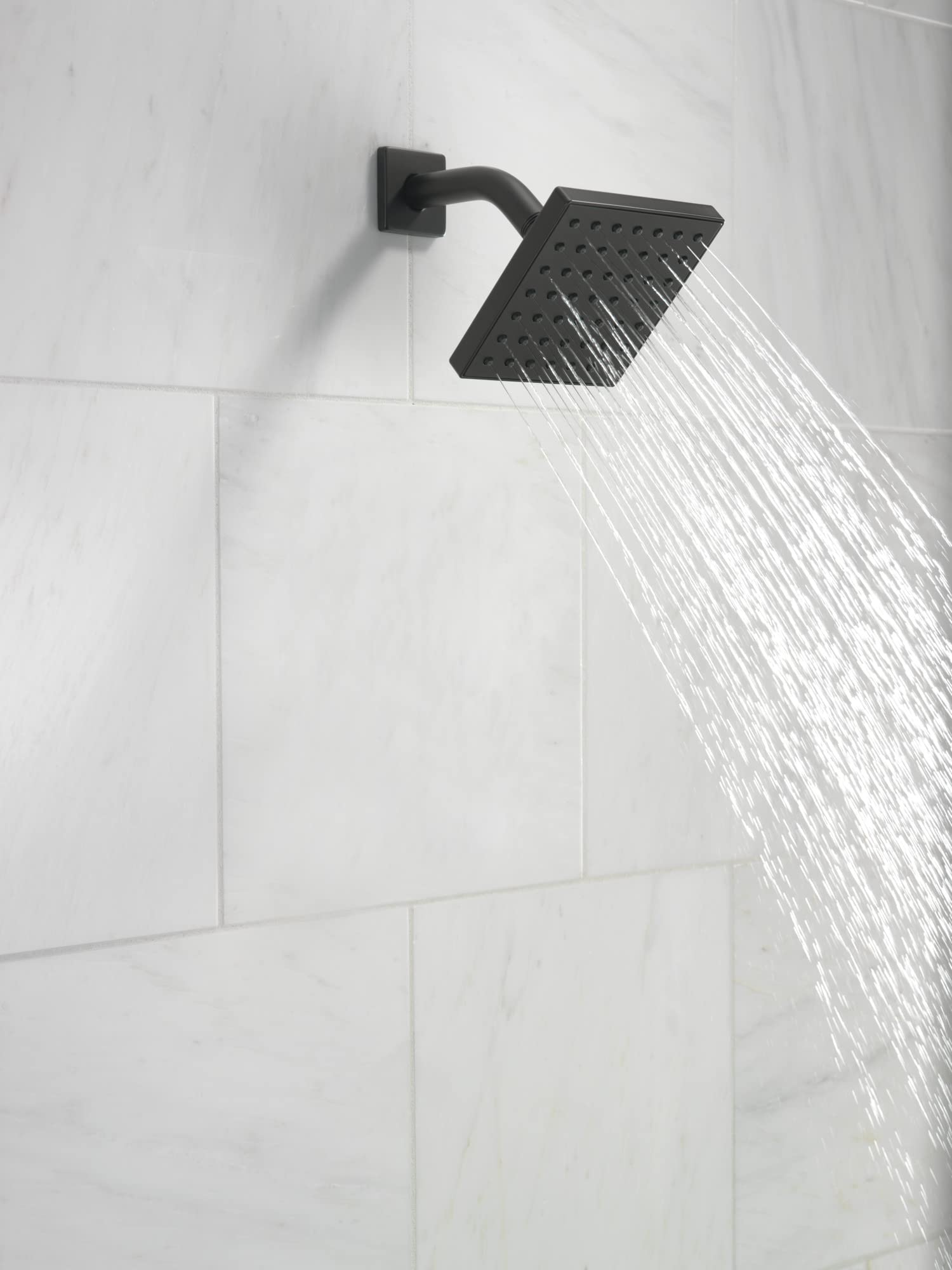 Matte black square shower head with curved arm mounted on white subway tile wall, water flowing from nozzles.