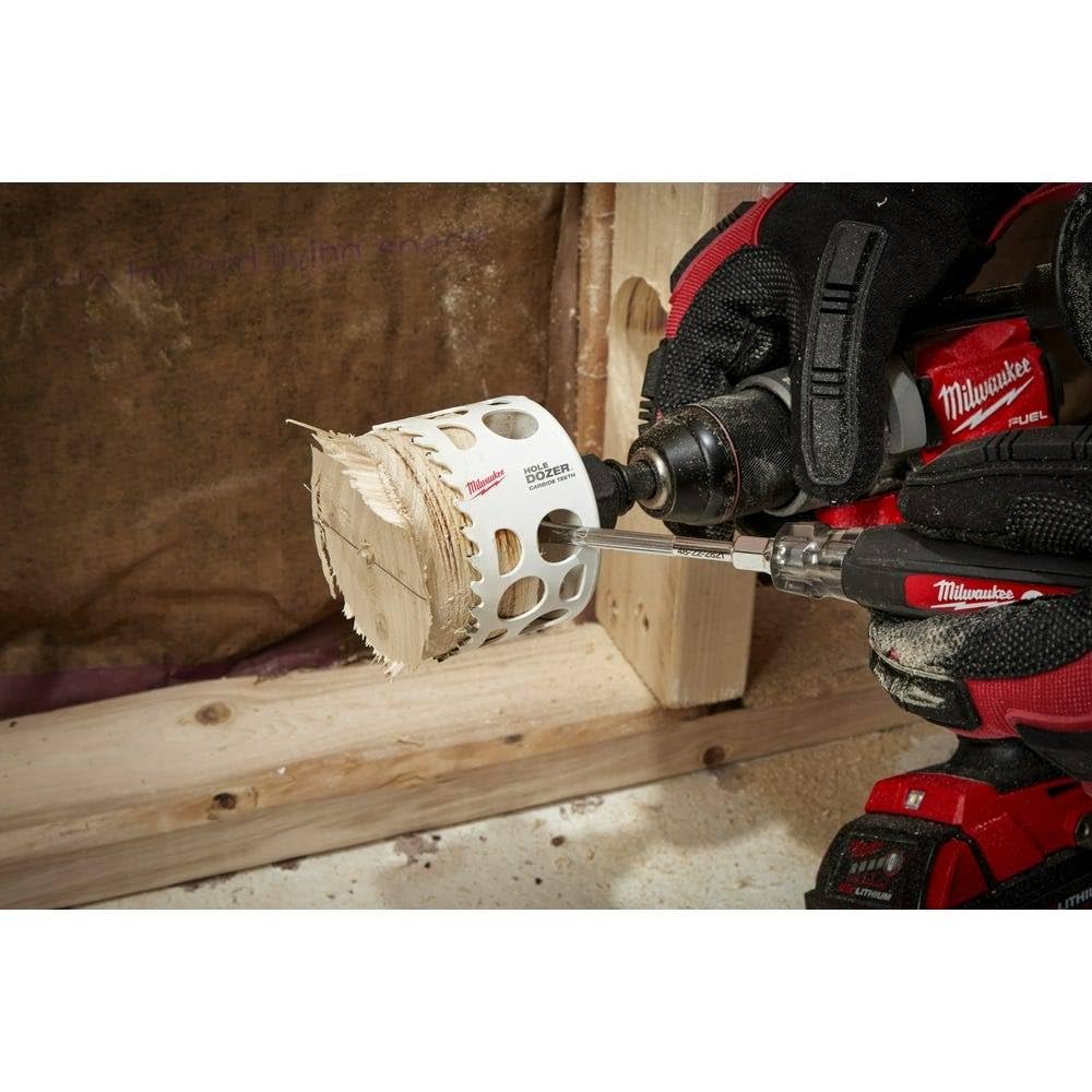 White cylindrical hole saw with carbide teeth and center pilot bit attached to Milwaukee drill in use on wooden surface.