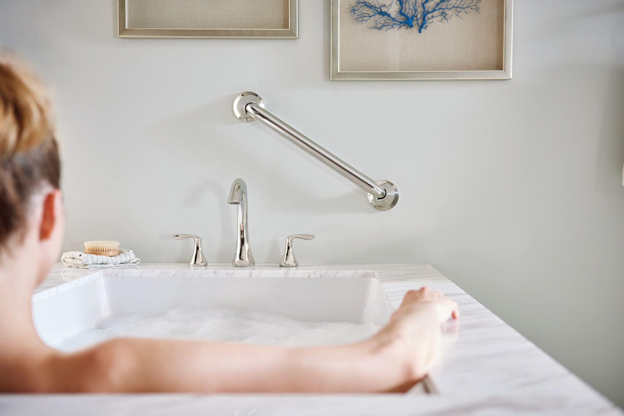 Chrome two-handle deck mount Roman tub faucet trim with curved spout and grab bar above white bathtub.