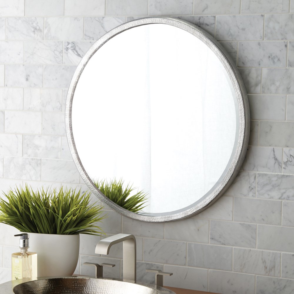 Round mirror with textured silver frame mounted on marble subway tile wall above bathroom sink.