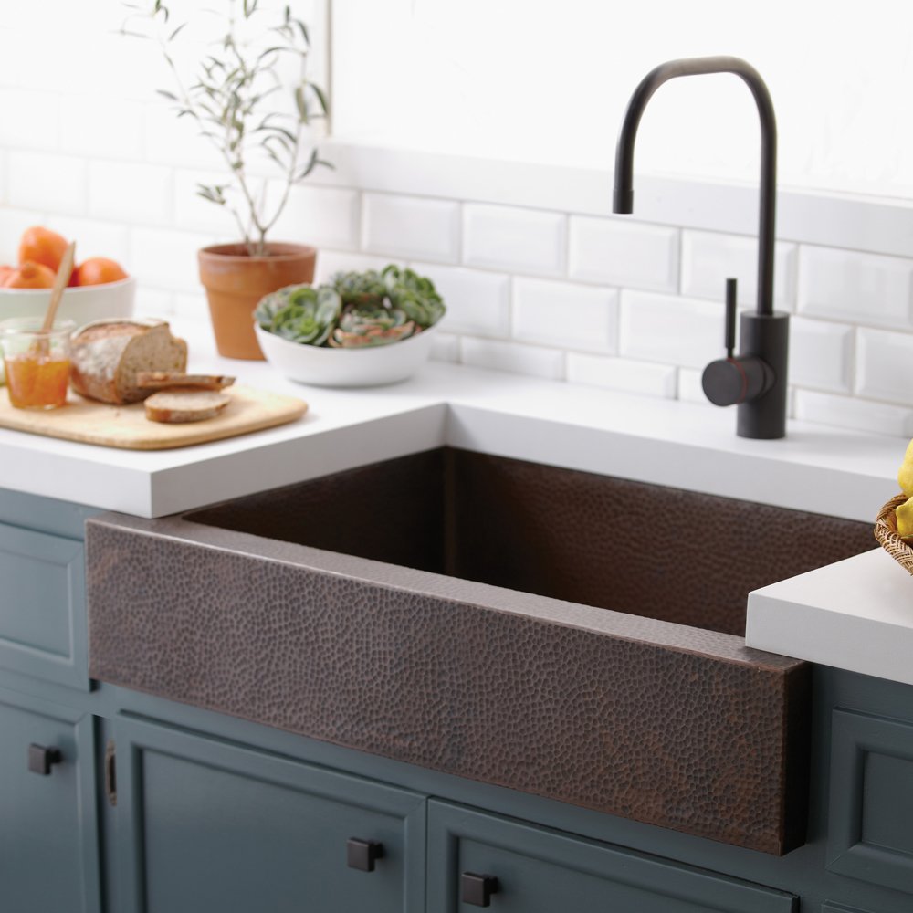 Antique copper farmhouse kitchen sink with hammered texture installed in white countertop with black faucet.