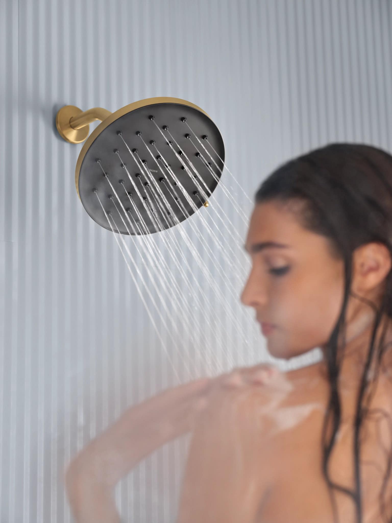 Brushed gold round rainshower head with multiple spray jets mounted on wall with water streams flowing downward.