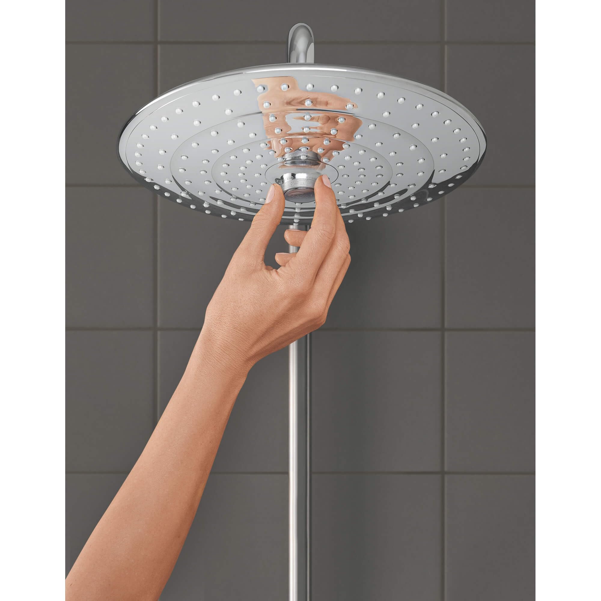 Chrome circular shower head with multiple spray holes held by a hand against tiled bathroom wall.