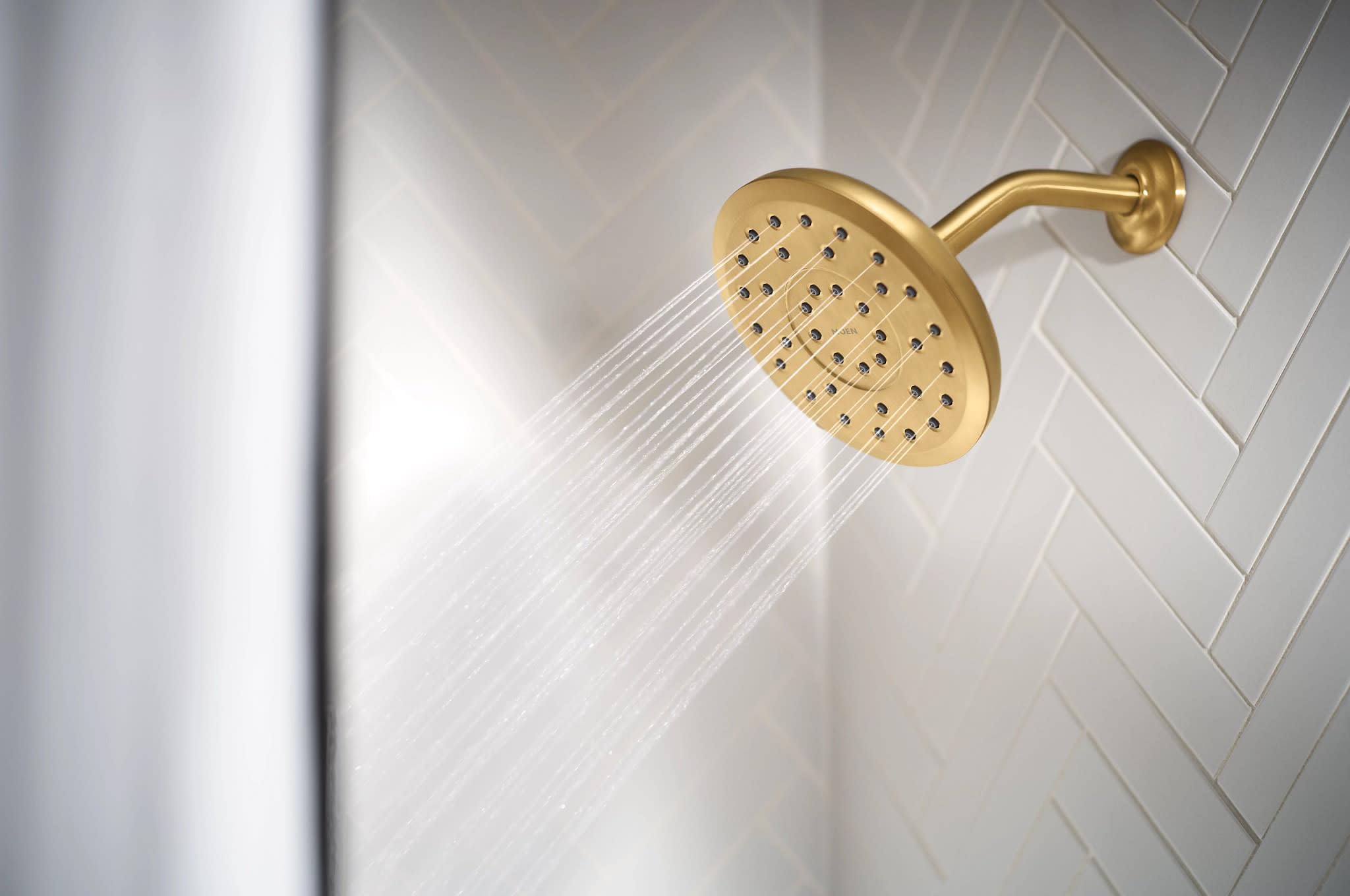 Round brushed gold shower head with multiple spray holes mounted on angled wall bracket with water streams visible.