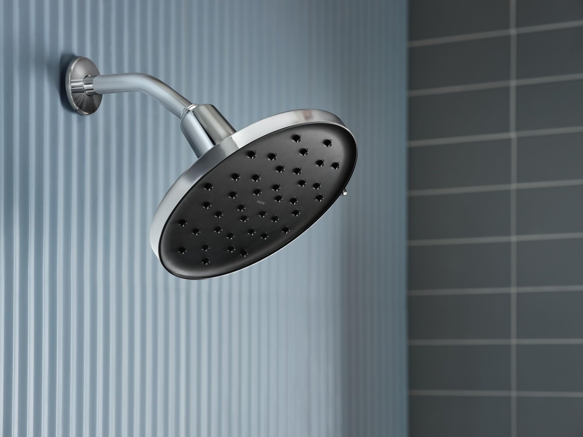 Chrome rainshower head with multiple spray holes mounted on curved arm against tiled bathroom wall.