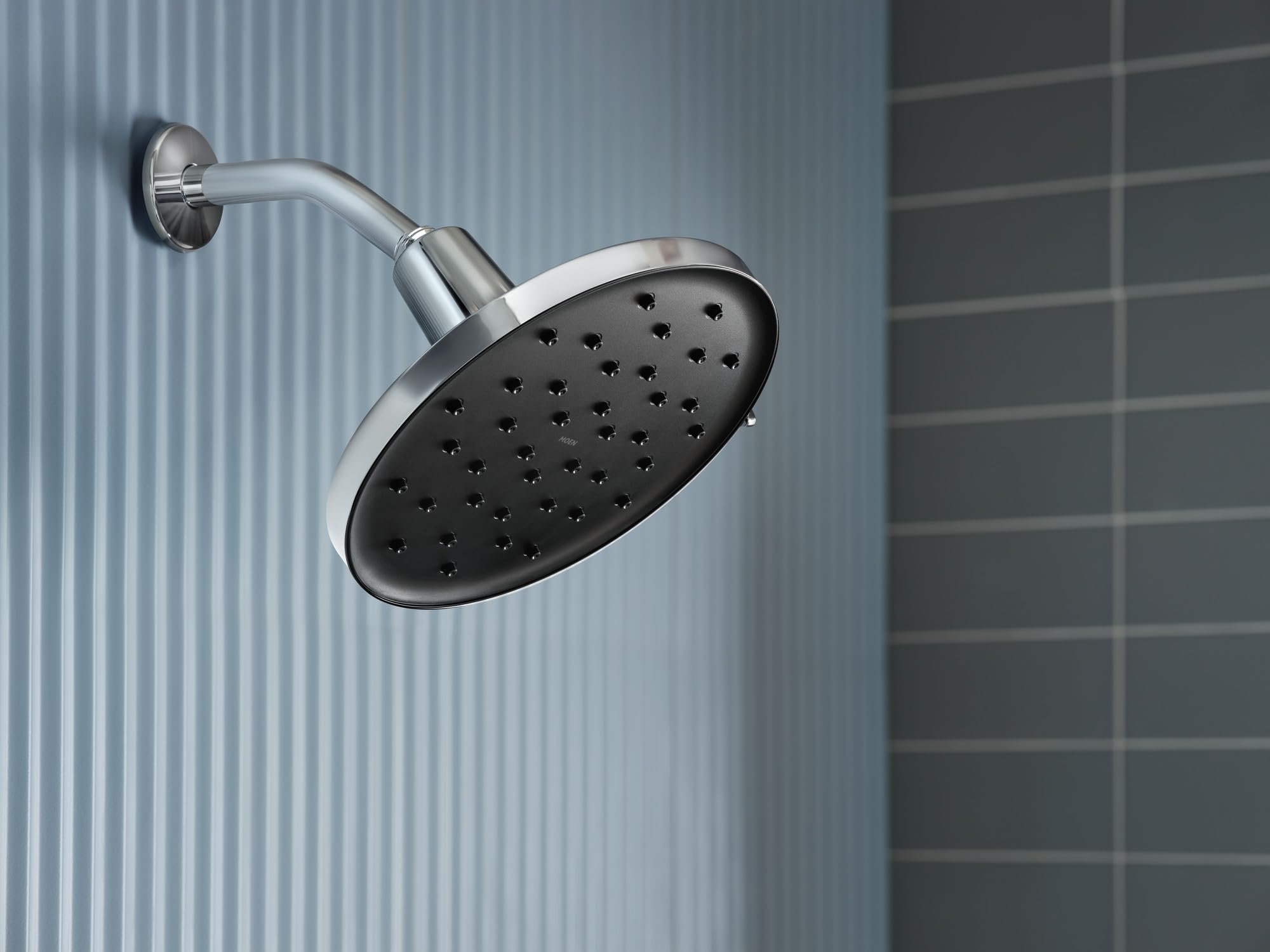 Chrome rainshower head with multiple spray holes mounted on curved arm against tiled bathroom wall.