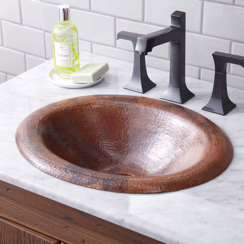 Oval copper bathroom sink with hammered finish set in marble countertop with black faucet and handles.