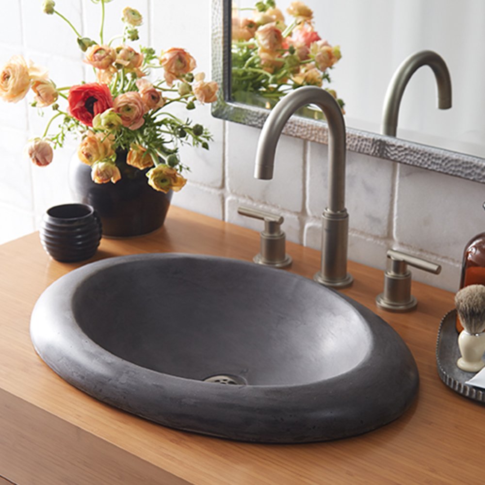Slate-colored oval bathroom sink with curved rim mounted on wood countertop with metal faucet and flowers.