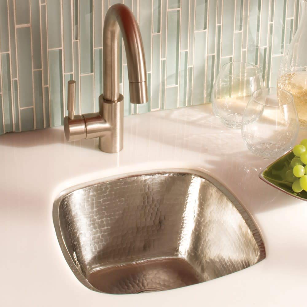 Small square hammered copper sink with curved sides and brushed nickel faucet on white countertop.