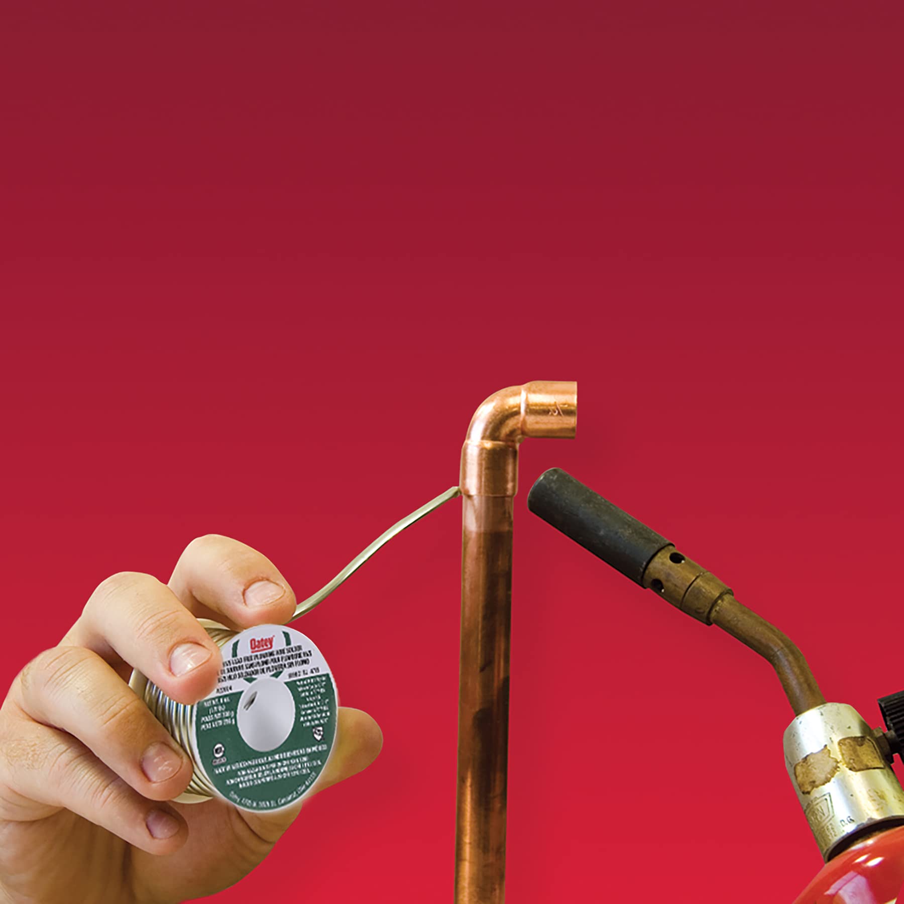 Hand holding gray solder wire spool next to copper pipe fitting and soldering iron against red background.