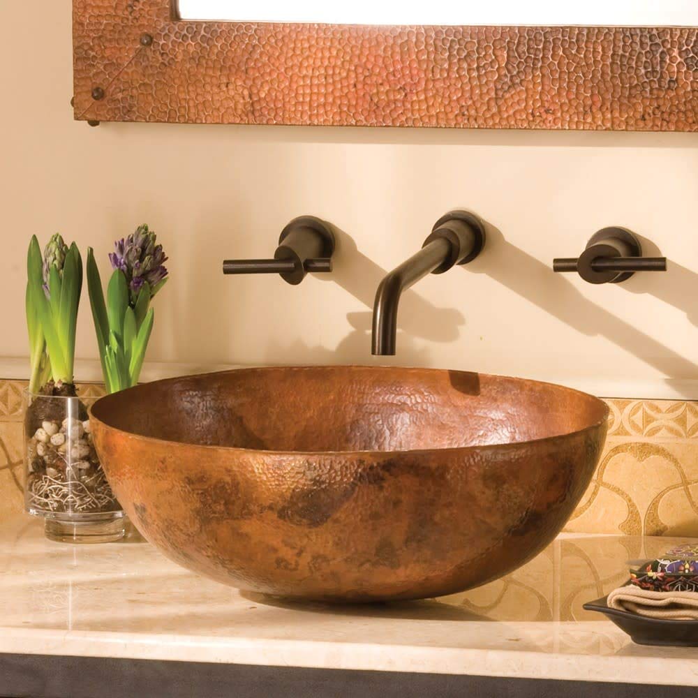 Oval copper bathroom sink with hammered antique finish and three wall-mounted bronze faucet handles.