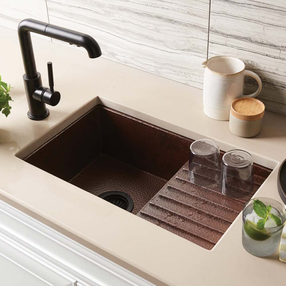 Hammered copper undermount bar sink with antique finish, featuring divided basin and drainboard, installed under white counte