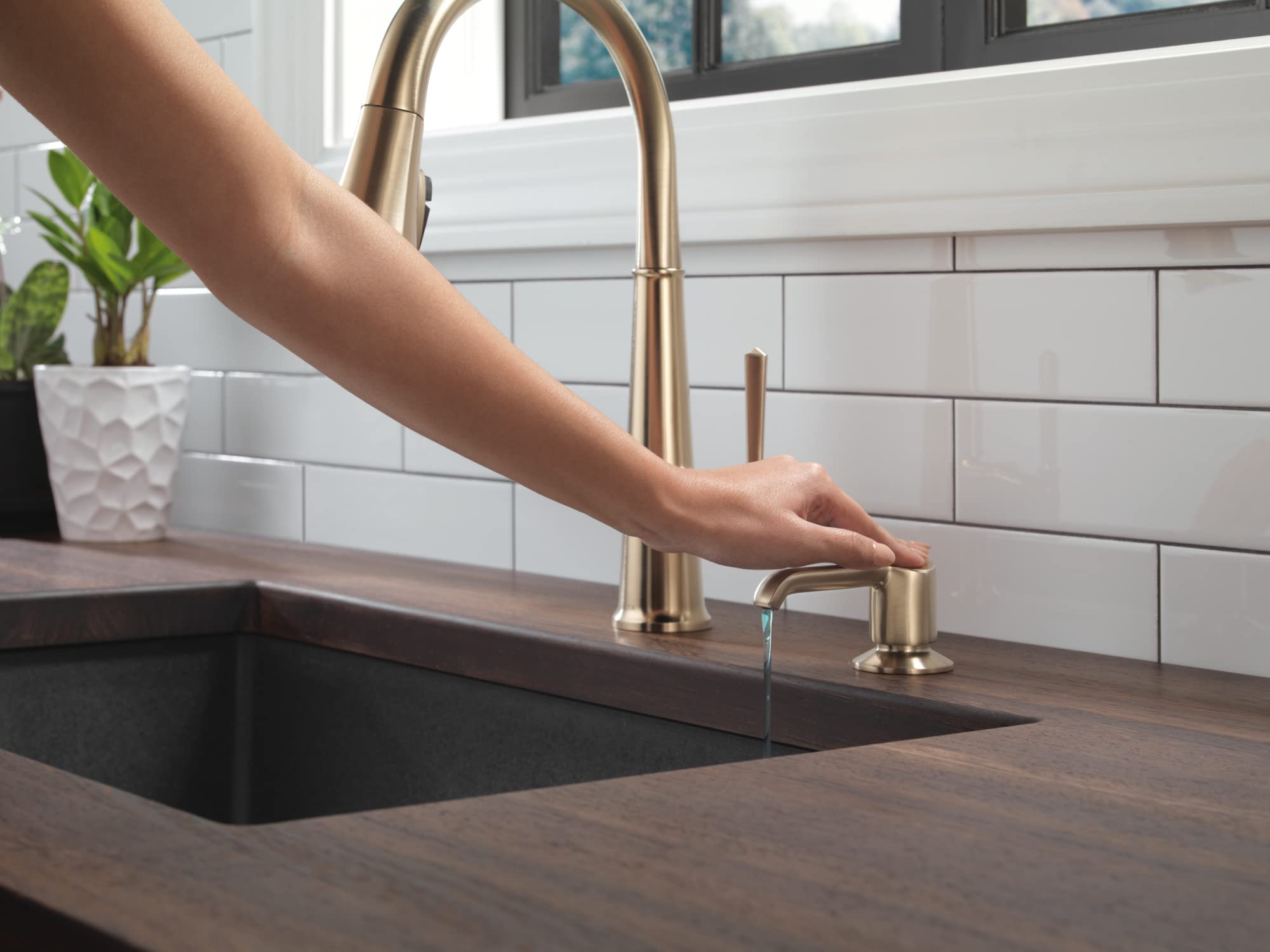 Champagne bronze soap dispenser with cylindrical pump next to kitchen sink faucet.