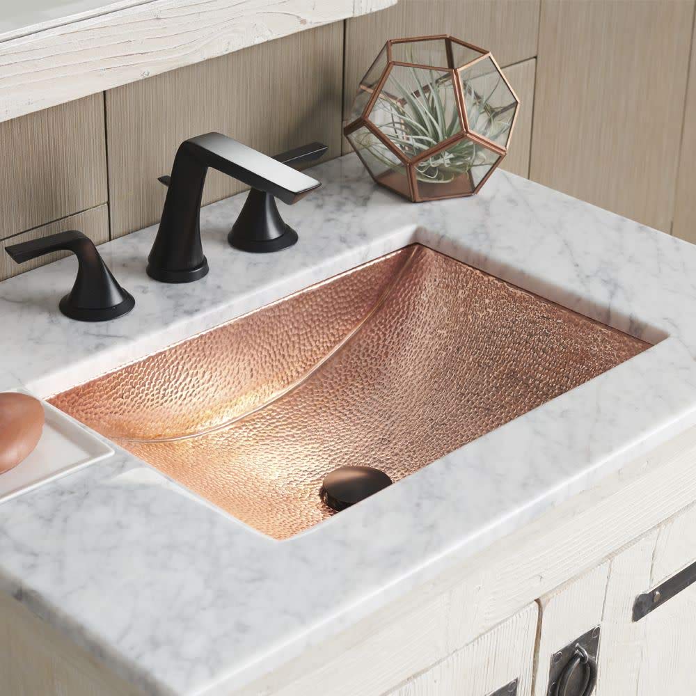 Polished copper rectangular undermount sink with hammered texture in marble countertop, paired with black faucet.