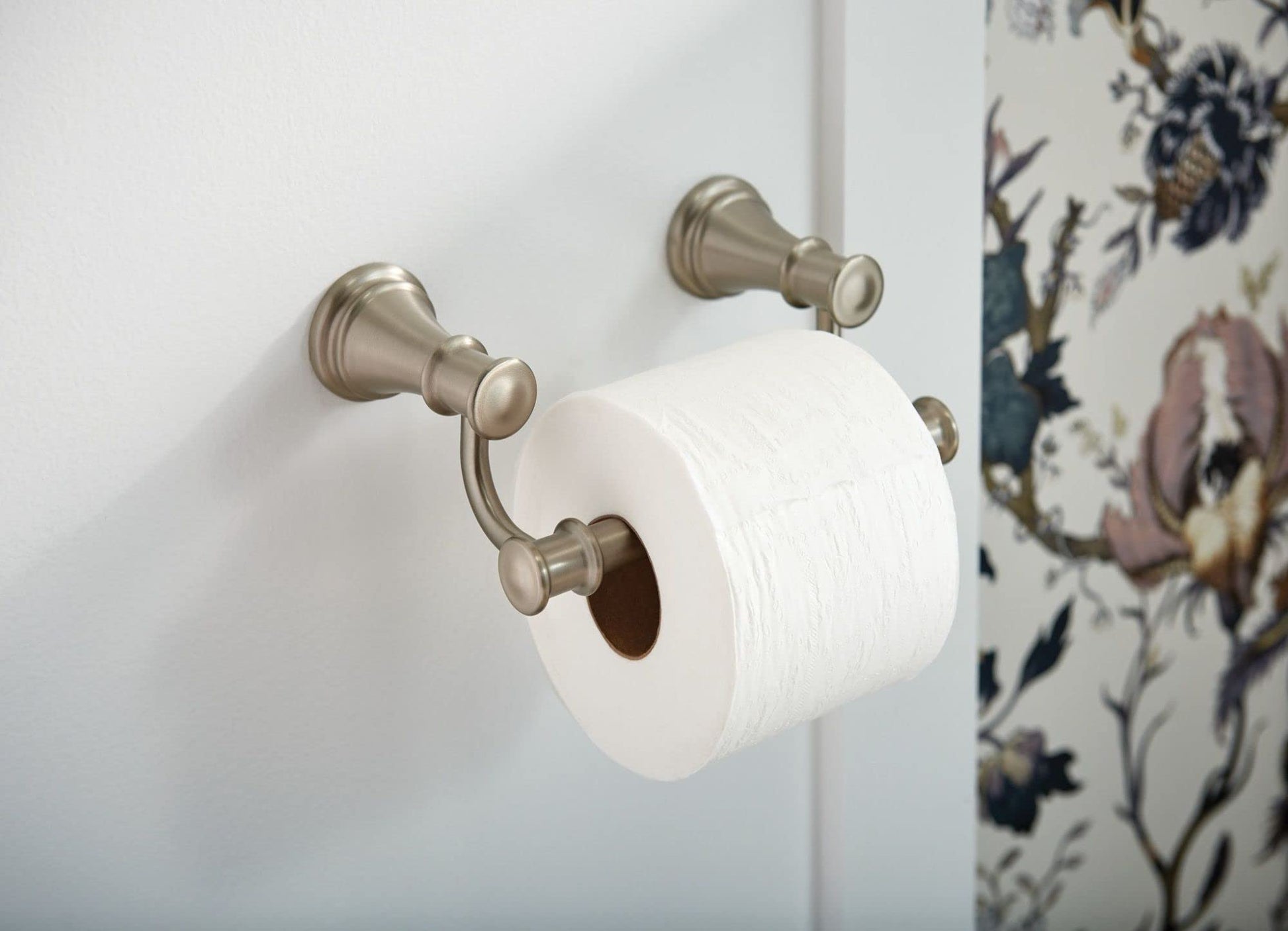 Brushed nickel toilet paper holder with decorative finials mounted on white wall with floral wallpaper.