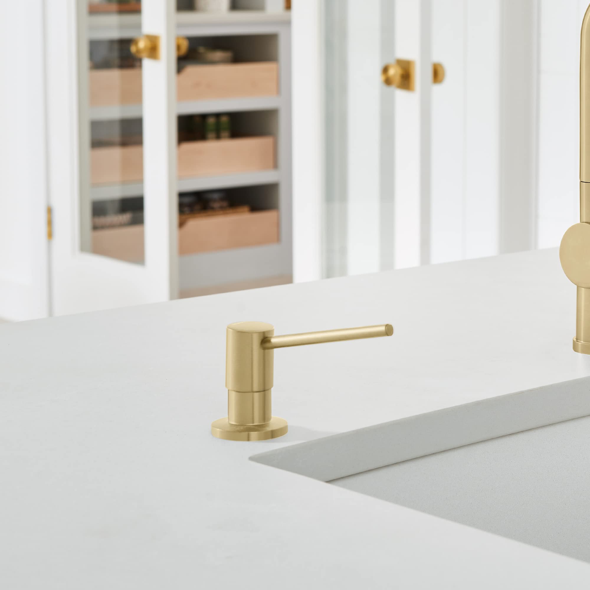 Satin gold soap dispenser with cylindrical pump mounted on white countertop in kitchen.