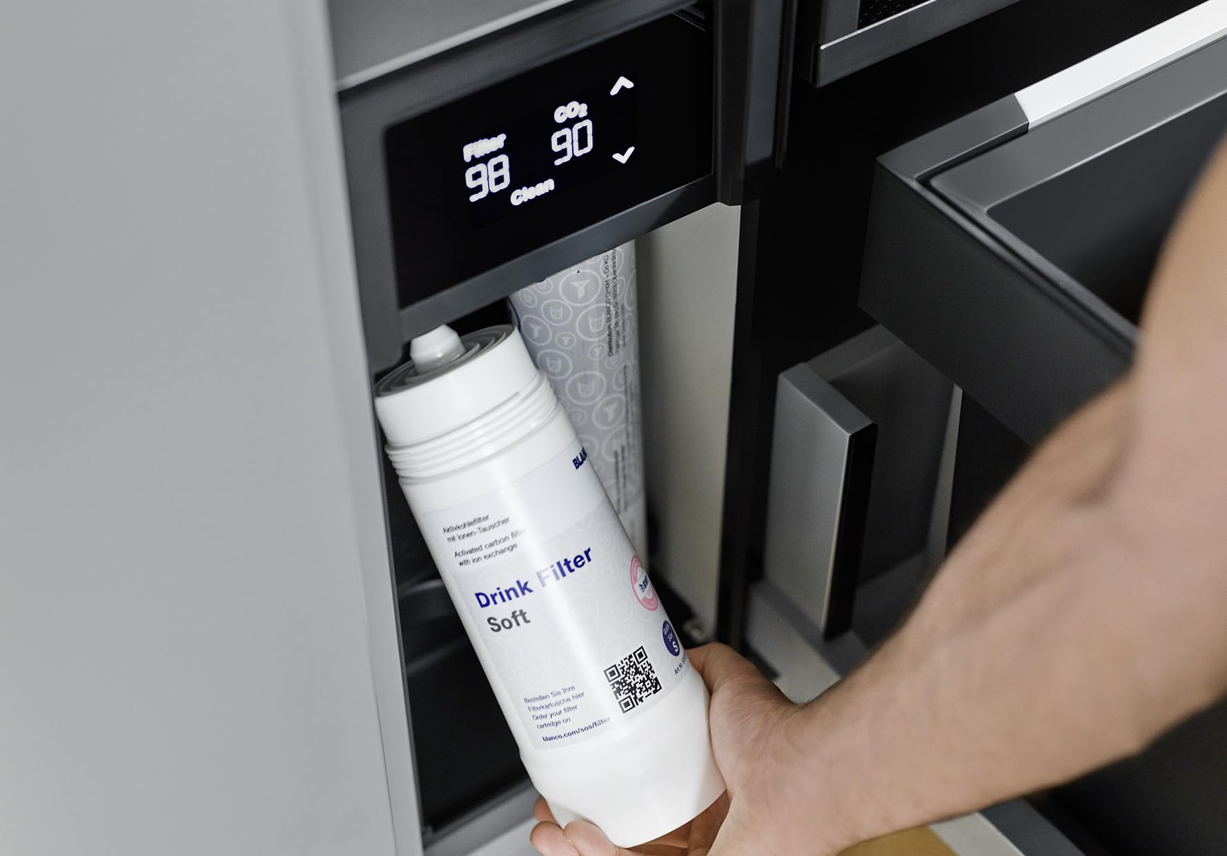 White cylindrical filter cartridge labeled "Drink Filter Soft" being held in front of a built-in kitchen water dispenser with