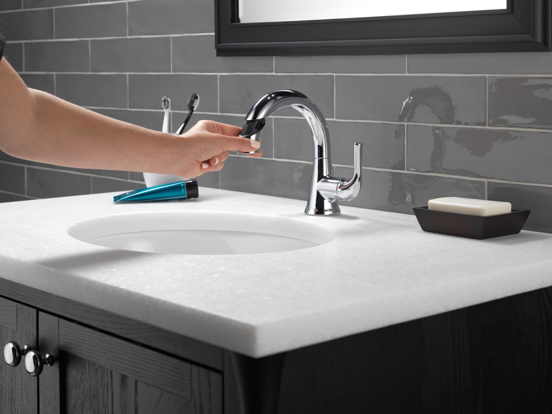 Chrome single-handle pull-down faucet installed on white bathroom sink with hand demonstrating spray function.