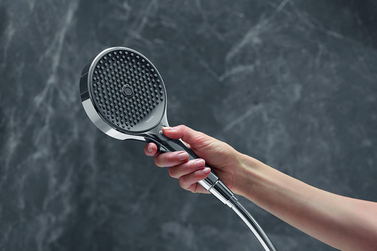 Chrome handheld showerhead with circular face and multiple spray holes, held in hand against tiled background.