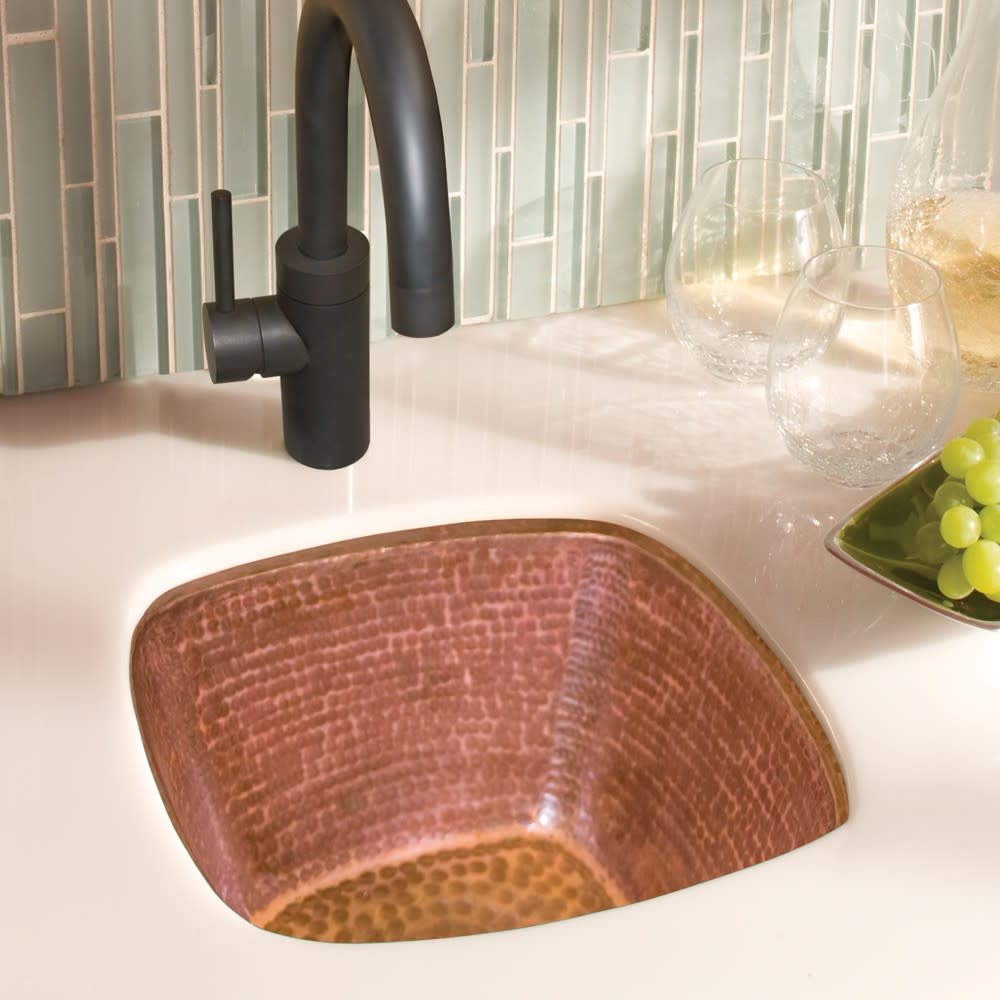 Small square hammered copper sink with textured surface installed in white countertop with black faucet.