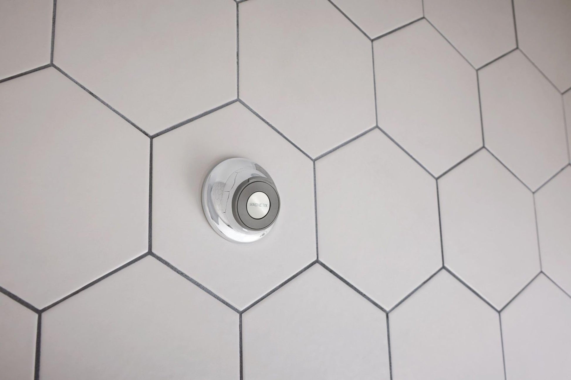 Circular magnetic cradle mounted on white hexagonal tile wall for holding showerhead remote.