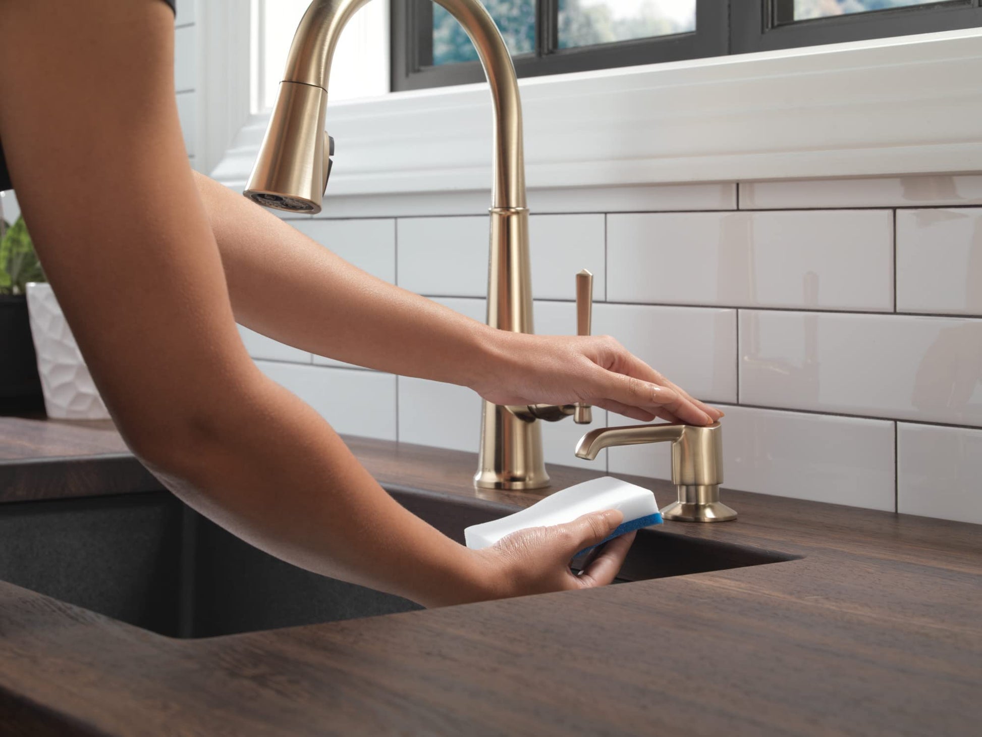 Champagne bronze soap dispenser with curved spout next to kitchen sink faucet, person's hand dispensing soap.