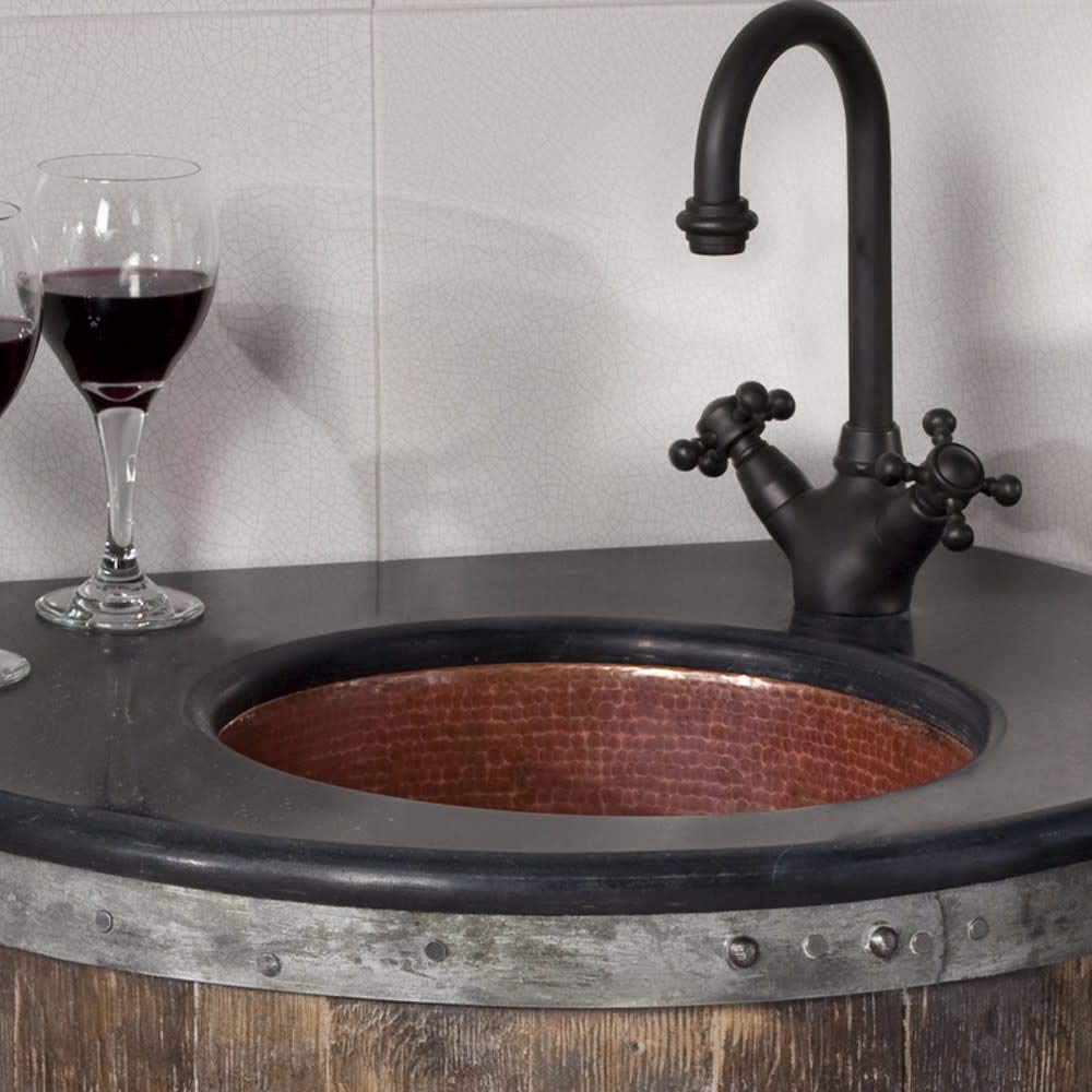 Hammered copper bar sink with dark rim mounted in countertop, paired with black gooseneck faucet and dual handles.