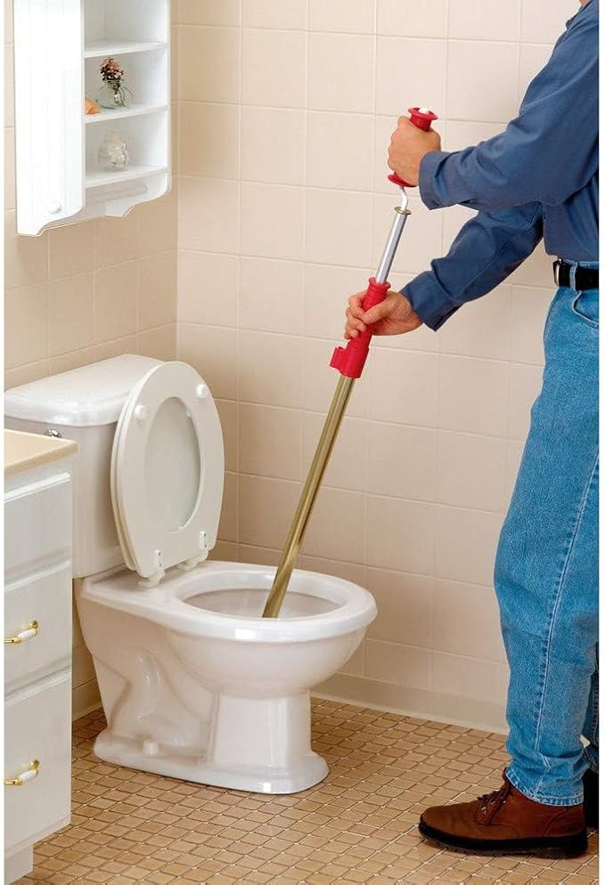 Person holding red-handled toilet snake auger with metal shaft positioned over white toilet bowl in bathroom.
