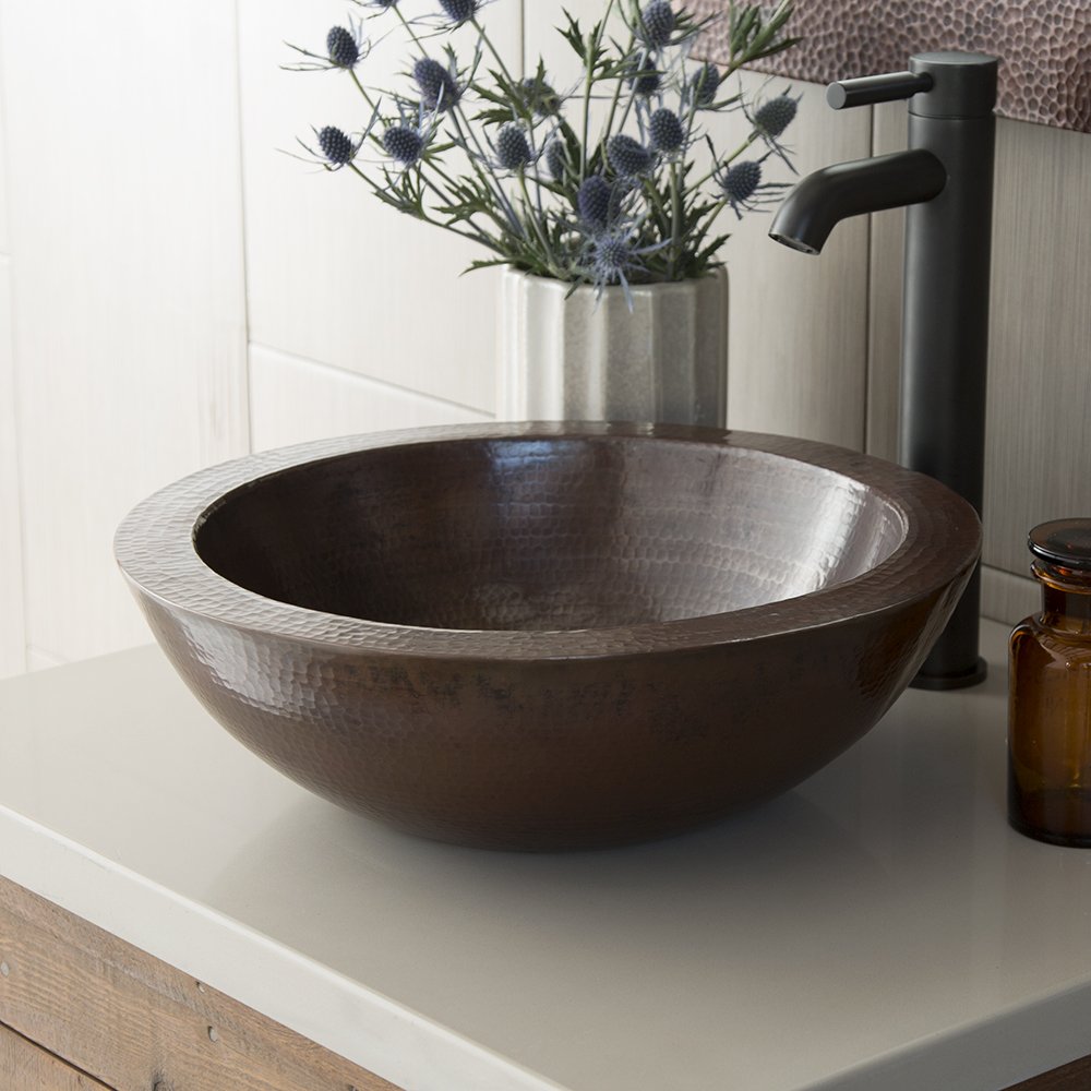 Round hammered copper vessel sink with antique dark finish on white countertop next to black faucet.