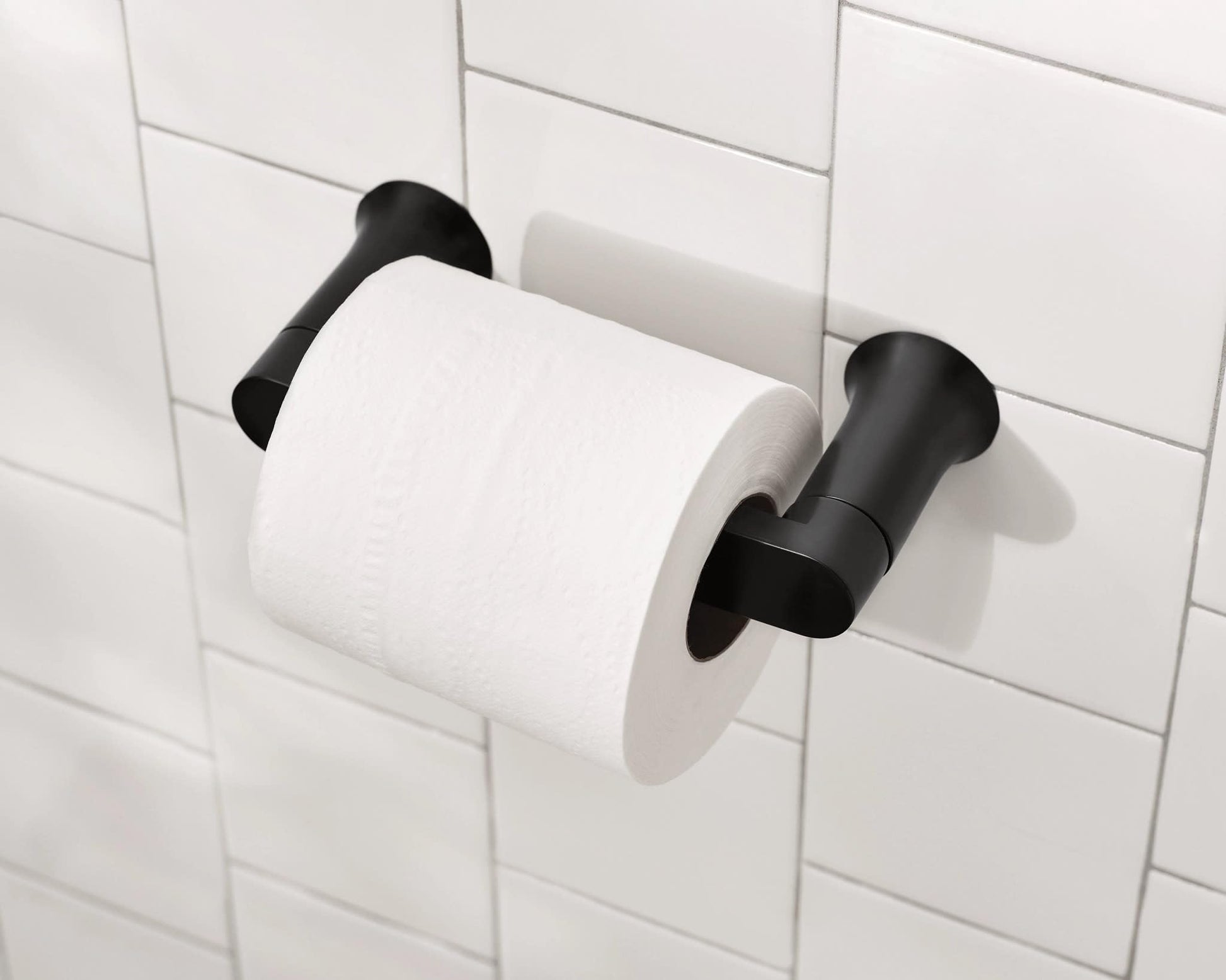 Black pivoting toilet paper holder mounted on white subway tile wall with white roll installed.