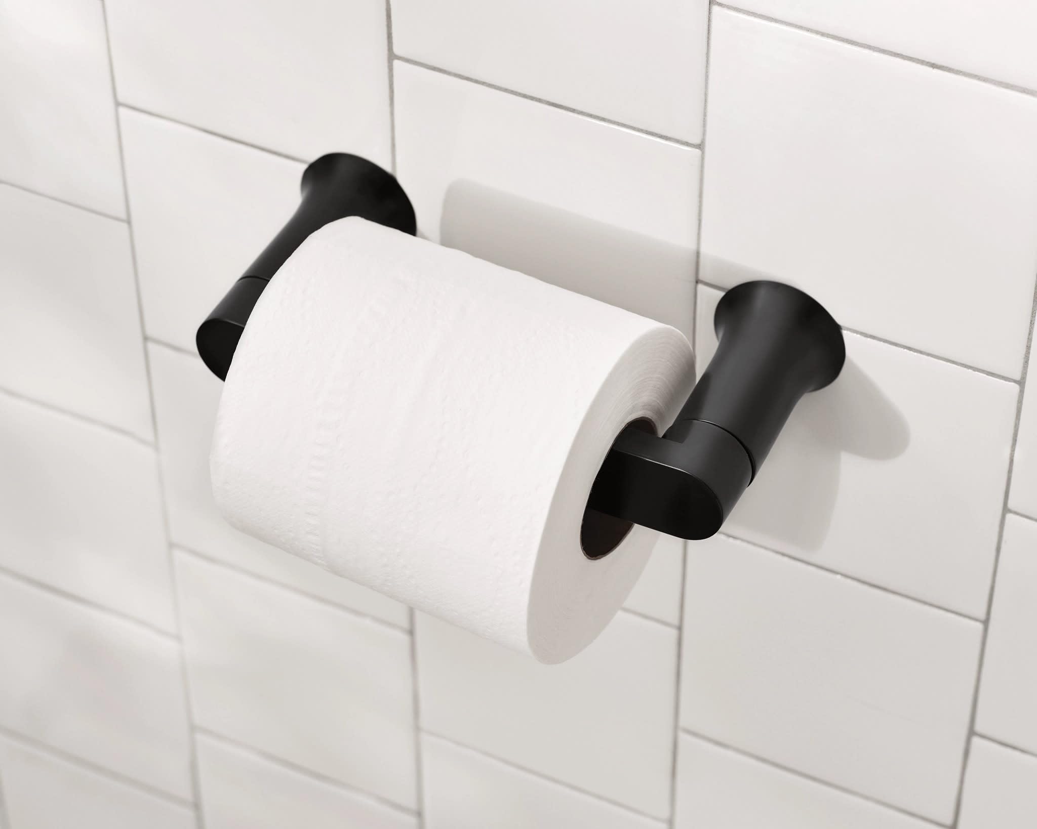 Black pivoting toilet paper holder mounted on white subway tile wall with white roll installed.