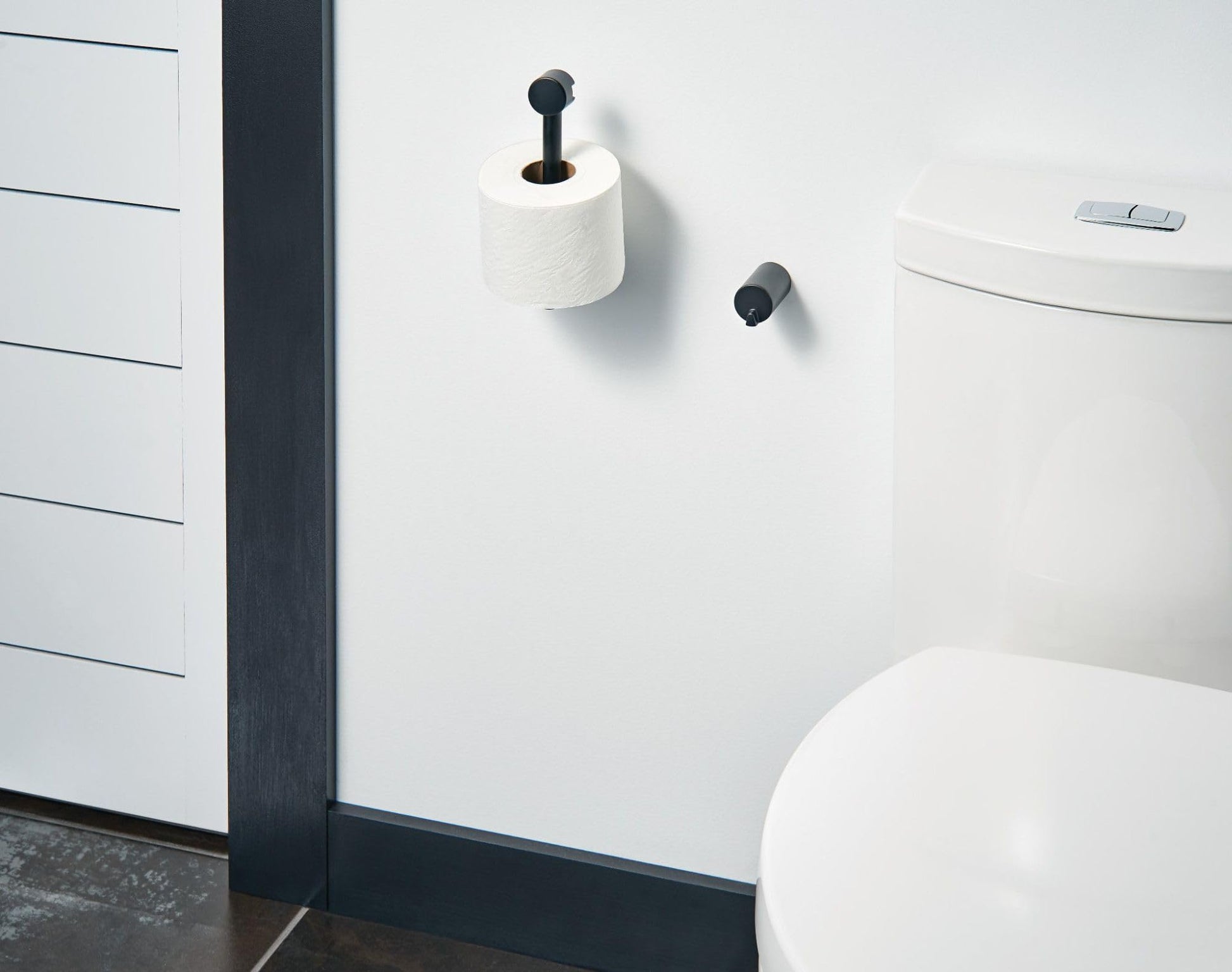 Black pivoting toilet paper holder mounted on white bathroom wall with roll installed.