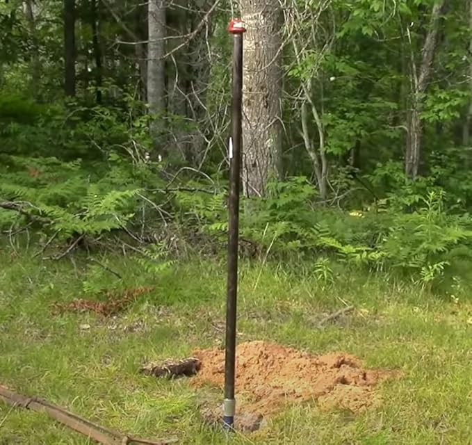 Stainless steel drive point with screen installed vertically in soil among trees and grass.