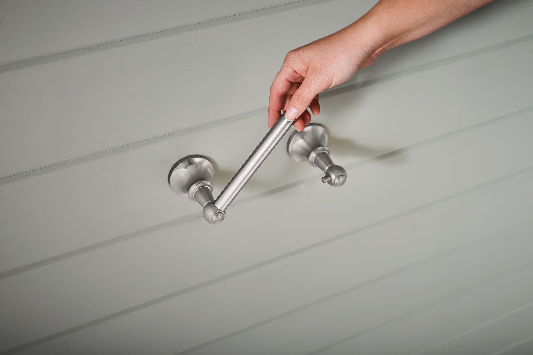 Brushed nickel pivoting toilet paper holder mounted on white wall with hand demonstrating pivot arm movement.