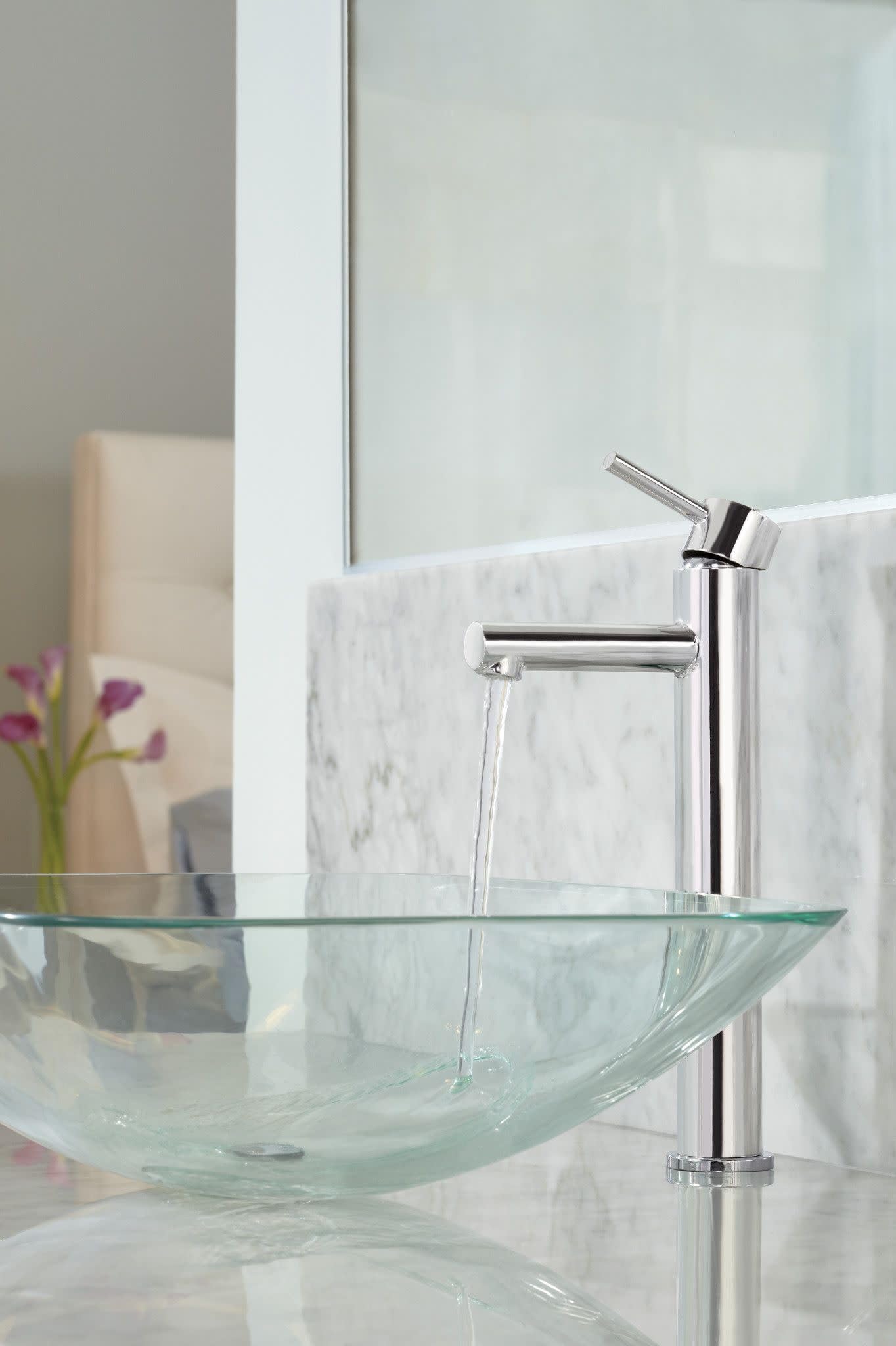 Chrome single-handle vessel sink faucet with tall spout mounted on marble backsplash above glass bowl sink.