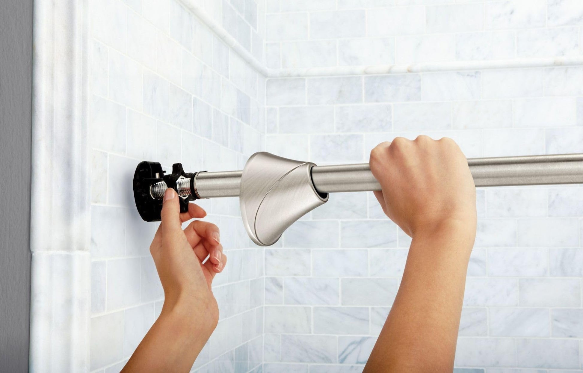 Brushed nickel curved tension shower rod being installed on white tile bathroom wall with hands adjusting brackets.