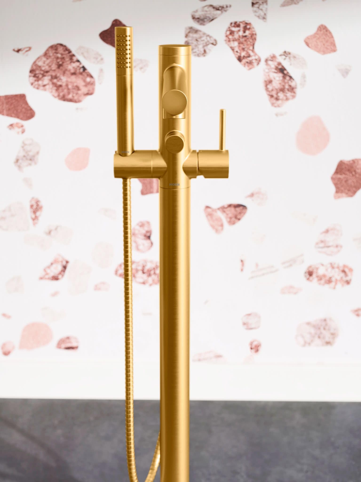 Brushed gold floor-mount tub filler with single handle, handshower on hose, and perforated showerhead.