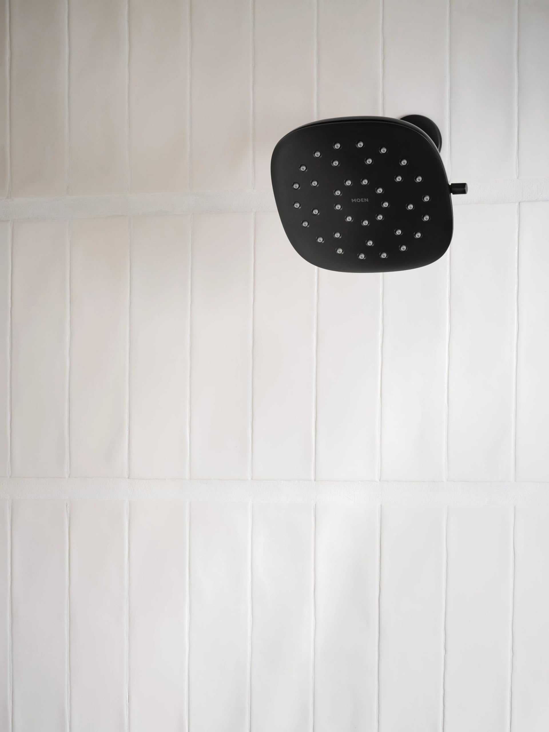 Matte black square rain shower head with multiple spray nozzles mounted on white subway tile wall.