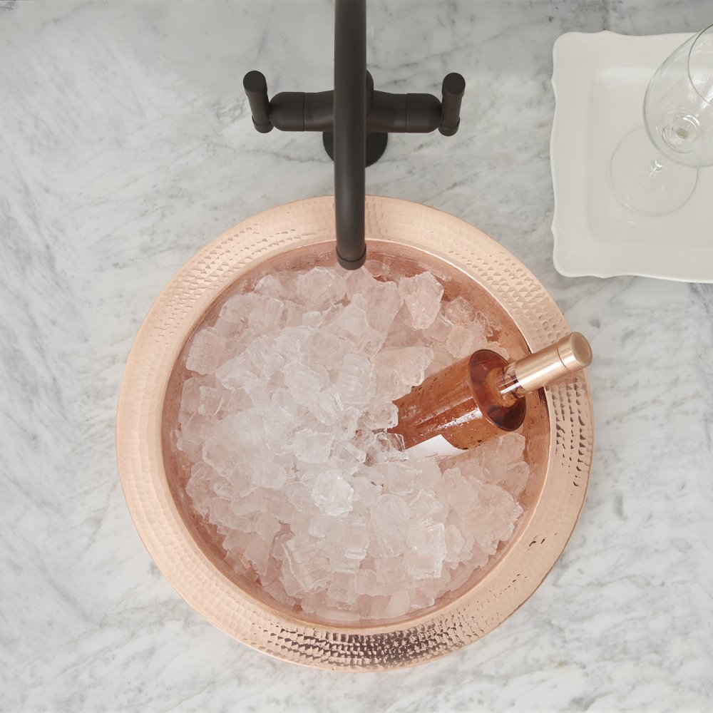 Polished copper round bar sink filled with ice and a muddler, paired with black faucet and handles, viewed from above.
