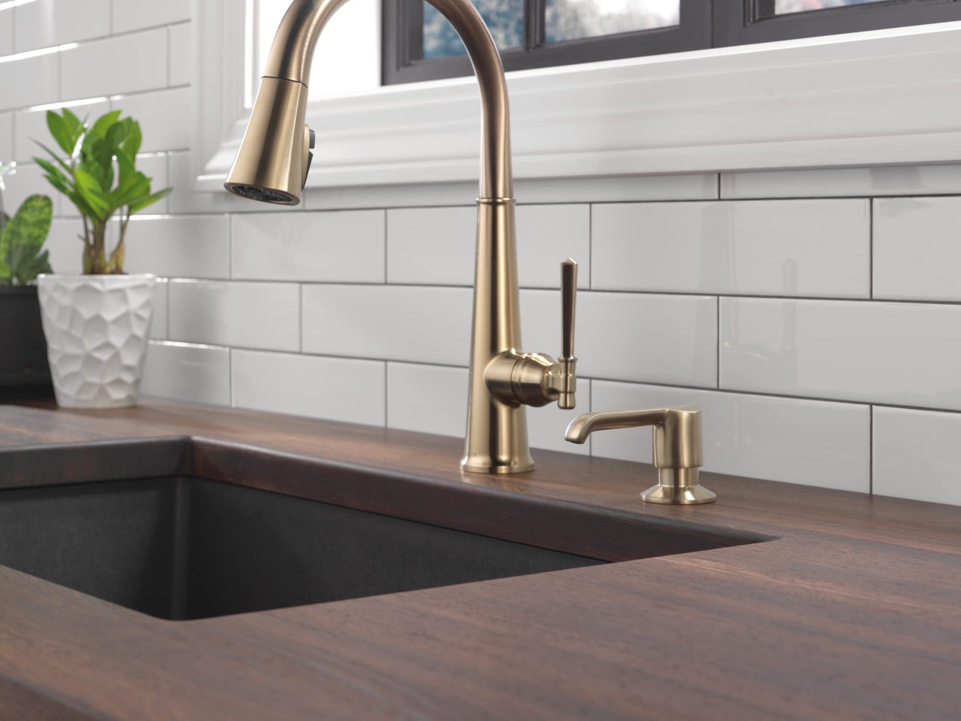 Champagne bronze soap dispenser with pull-down spray faucet mounted next to kitchen sink with white tile backsplash.