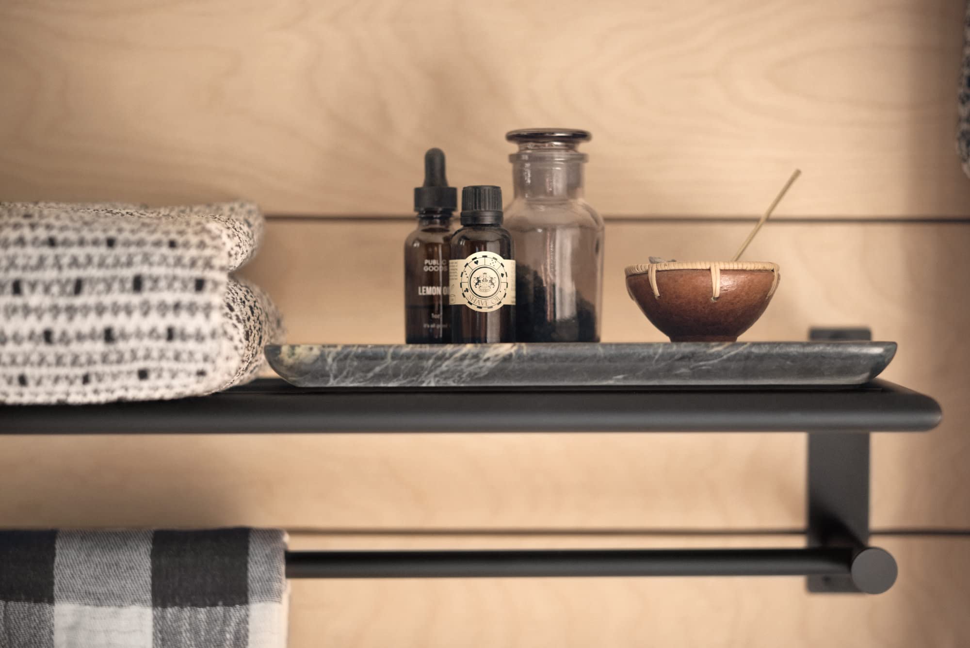 Matte black bathroom shelf with towel bar holding folded towels, bottles, and bathroom accessories.