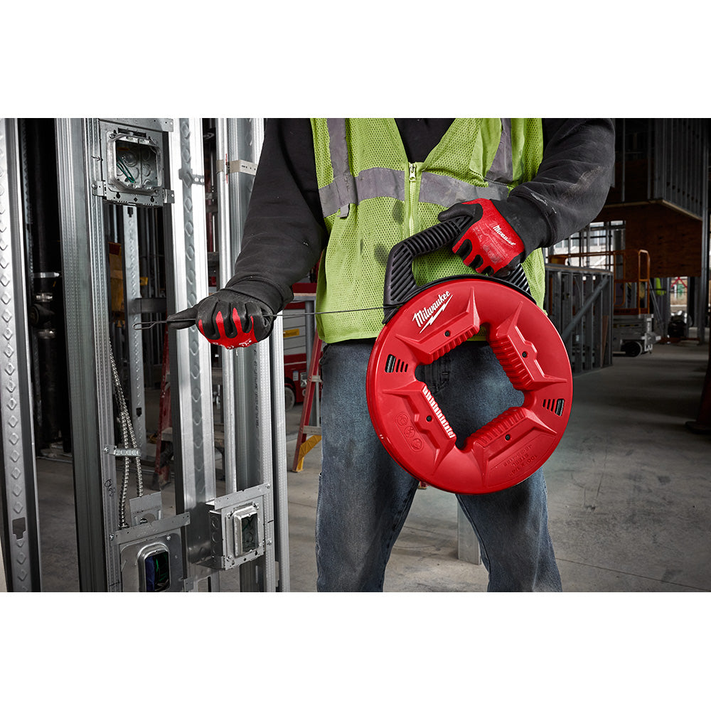 Worker in safety gear holding red Milwaukee fish tape reel with steel tape extended toward electrical conduit.