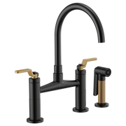 Black bridge faucet with curved arc spout, gold lever handles, and matching side spray with textured grip.