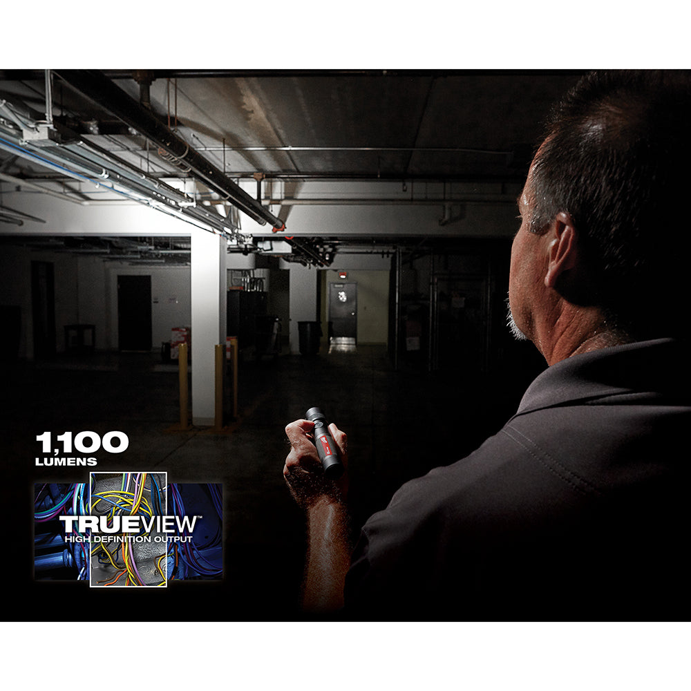 Man holding cylindrical rechargeable flashlight illuminating a dark industrial warehouse interior.