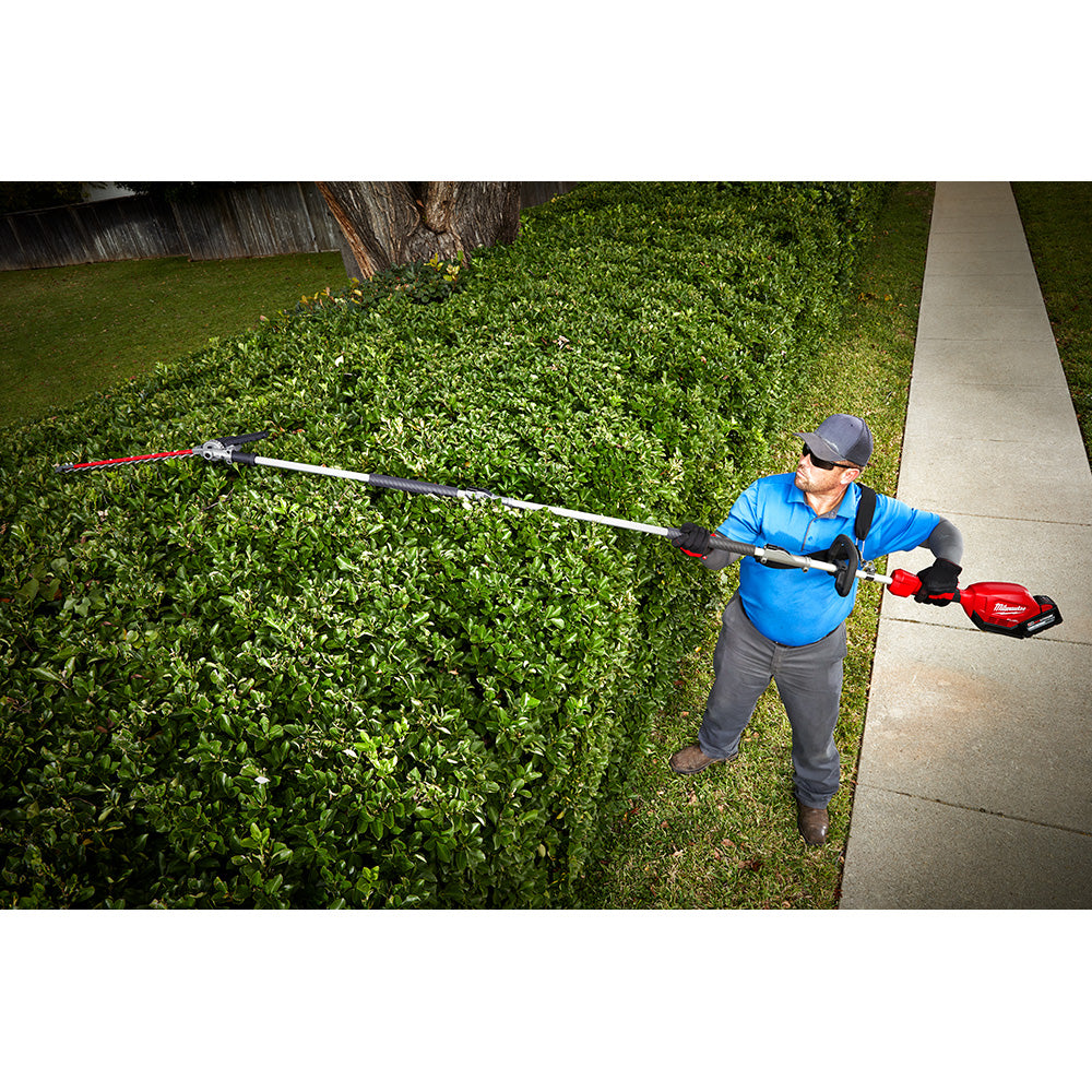 Person using extended hedge trimmer attachment on tall green shrub in residential yard.