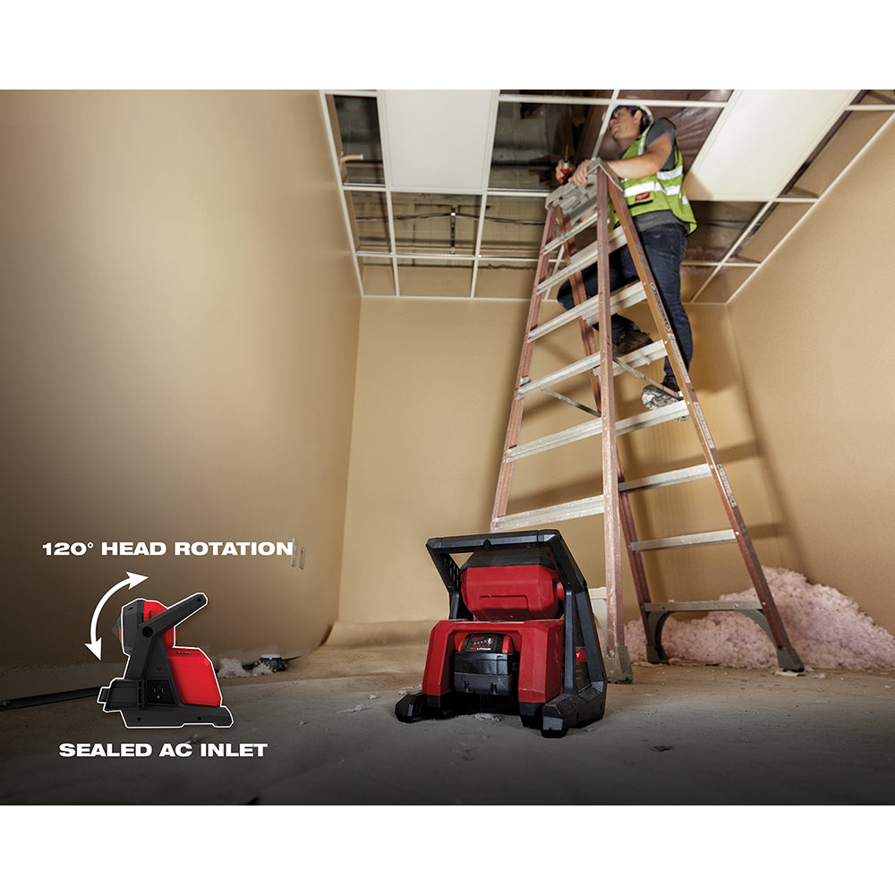Red and black Milwaukee M18 flood light positioned on a basement floor with a worker on a ladder above.