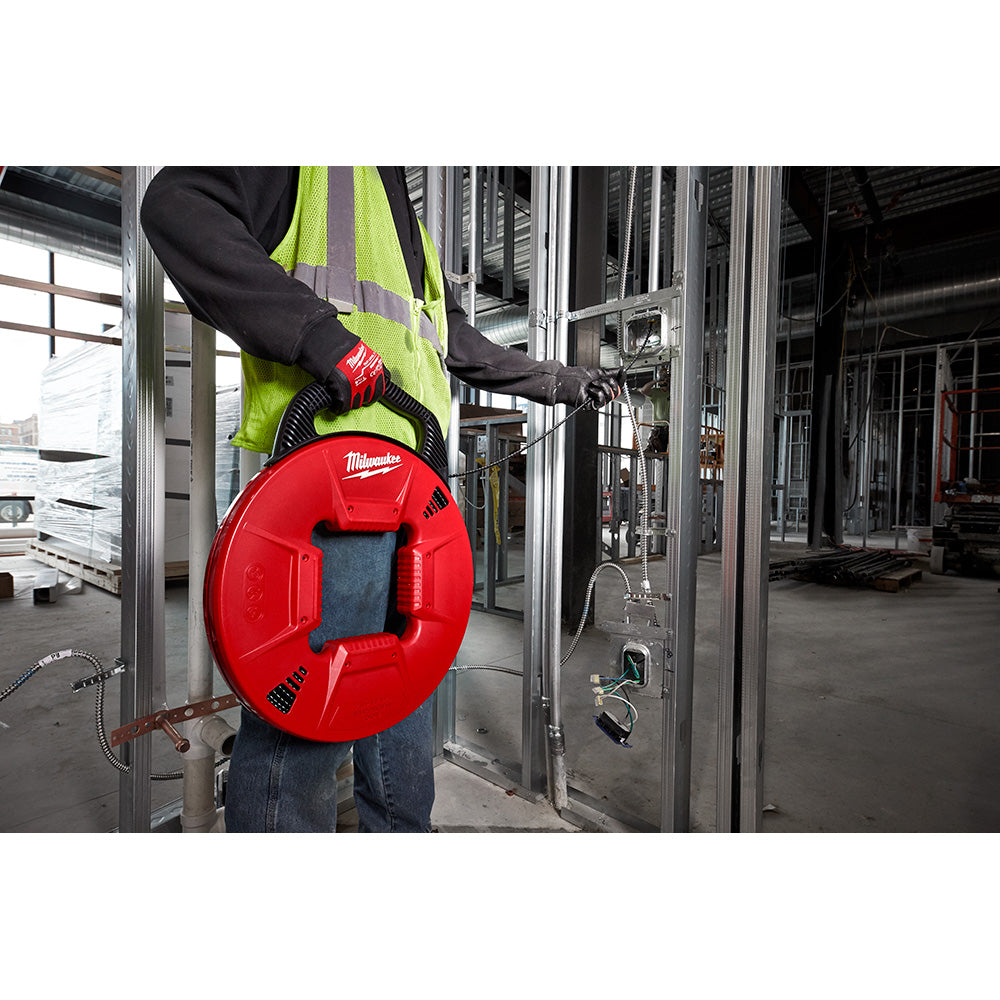 Worker in safety vest holding red Milwaukee fish tape reel with metal leader extending into electrical conduit.
