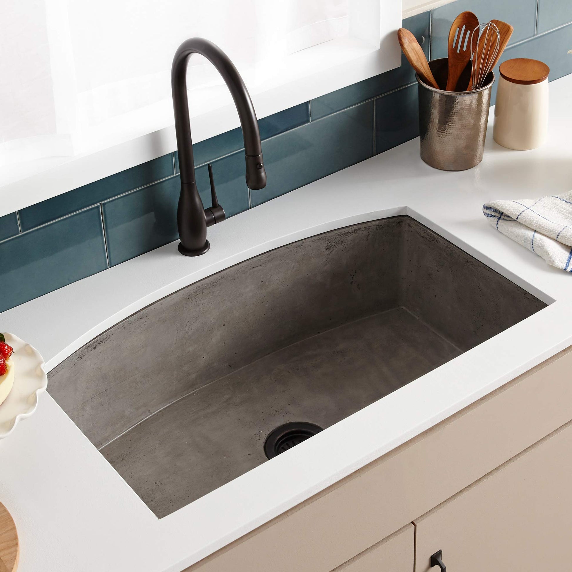 Ash concrete farmhouse kitchen sink with single black faucet, installed in white countertop with teal tile backsplash.