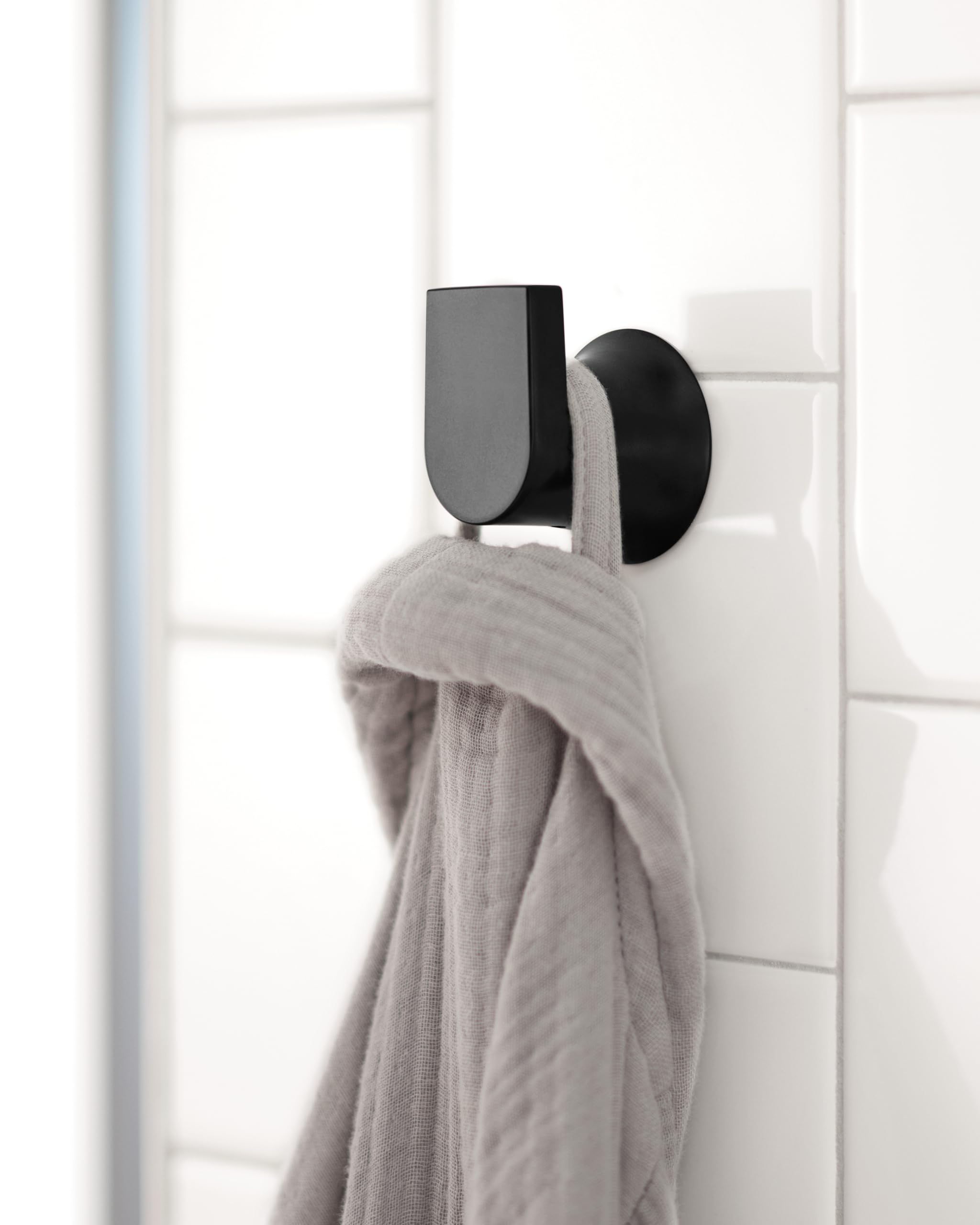 Black wall-mounted robe hook with rounded design holding a gray towel on white tile wall.