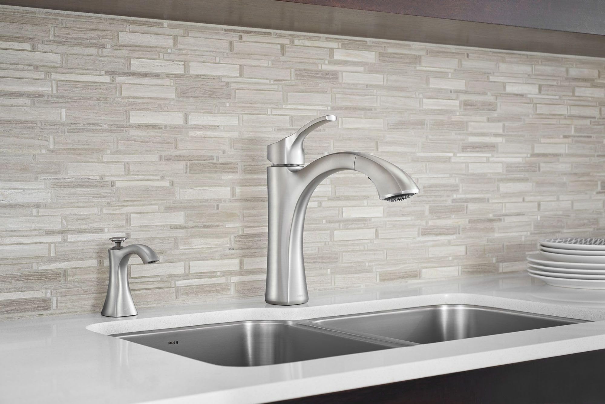 Chrome soap dispenser and kitchen faucet installed on countertop next to stainless steel sink.